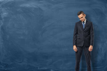 A sad businessman in a suit standing with bowed head and his arms down near the blue chalkboard. Face serious challenges. Business difficulties. Get into trouble.の写真素材