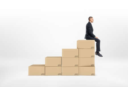 A businessman sitting on top of closed cardboard boxes put together in the shape of stairs, isolated on a white background. Postal services. Packing and crating. Business development. Company manager.の写真素材