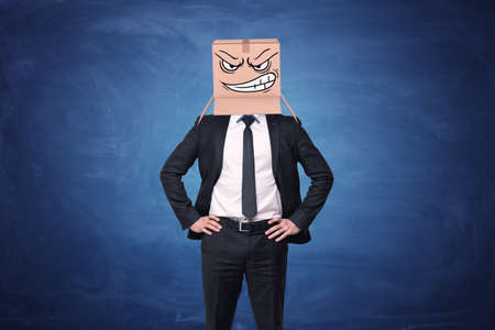 Businessman is wearing cardboard box with drawn angry face on his head on blue chalkboard background . Anonymous person. Aggressive marketing strategies. Business issues.の写真素材