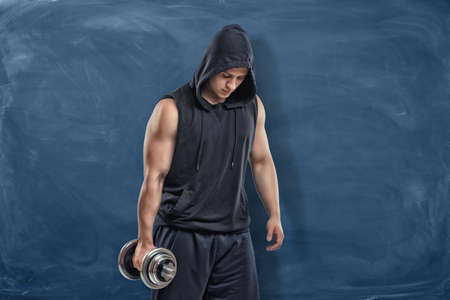 Portrait of a young handsome man in a black outfit standing and holding a silver dumbbell in his hand during training on blue chalkboard background. Sport and healthy lifestyle. Keep fit. Athletic body. Exercises and warming up.の写真素材