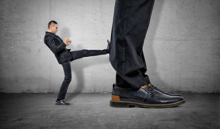 Tiny businessman kicking huge legs of another on the gray concrete background. Business rivalry. Confident behaviour. Career growth.の写真素材