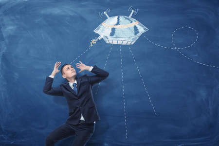 Businessman frightened by flying saucer painted on blue blackboard behind himの写真素材