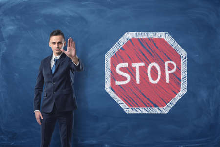 Businessman standing with his arm forward and traffic-sign Stop painted on blackboard behind himの写真素材