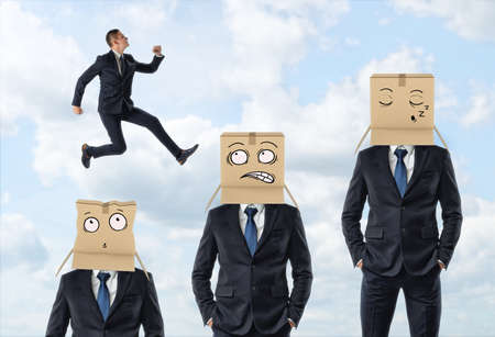 Small businessman jumping over three men in suits who are wearing carton boxes with painted faces.