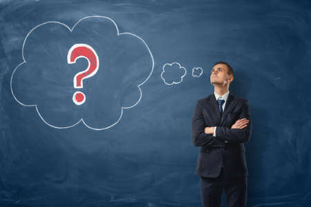 Businessman on blue blackboard background where thoughts bubbles with a red question mark are drawn in chalk.の写真素材
