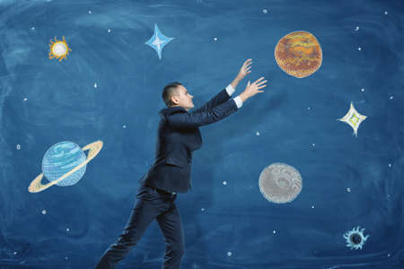 Businessman on blue chalkboard background trying to grasp a drawn planet among many others.の写真素材