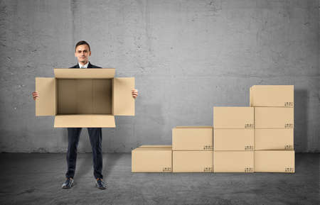 Businessman is showing an open box in his hands staying on gray background