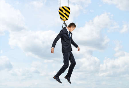 Businessman is being lifted by a crane hook on the background of sky and cloudsの写真素材