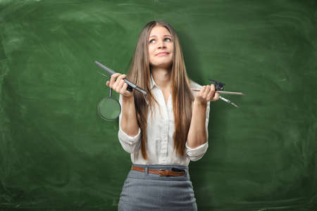 Confused businesswoman on green chalkboard background holding pens, ruler and magnifying glass in her hands.の写真素材