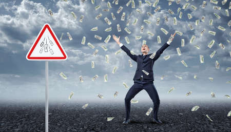 Ecstatic businessman standing under many dollar bills falling from the sky with a traffic warning sign Money nearby.の写真素材
