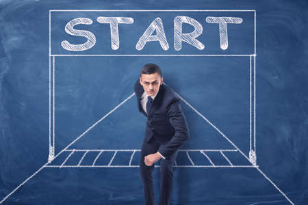 Businessman standing in running position on blue blackboard background with chalk drawing of starting gate.の写真素材