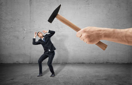 Scared businessman full-height in a desperate pose and a giant hand with a hammer threting him on the gray backgroundの写真素材