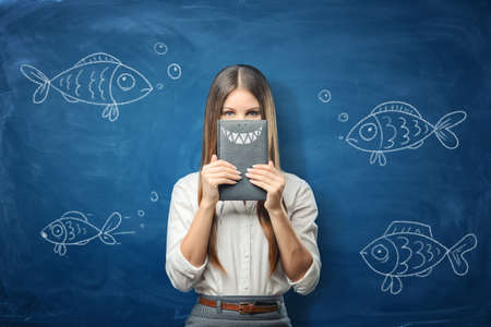 Woman is holding a notebook with drawn shark teeth on blue chalkboard background with drawn fishの写真素材
