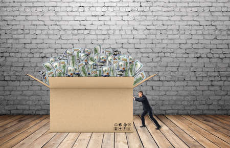 Businessman is pulling a huge box of money in front of him on the background of brick wall and wooden floorの写真素材