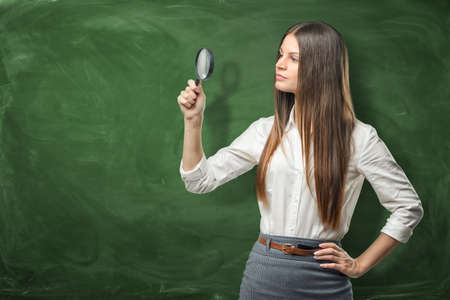 Young woman in official clothing is looking with a magnifying glass on the background of green blackboardの写真素材