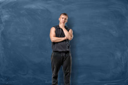 Muscular young man is standing and thinking holding his head with hand on blue chalkboard backgroundの写真素材