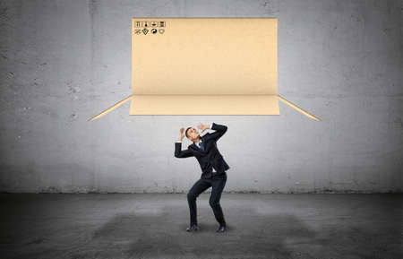 Businessman on concrete background cowering below a large open carton box falling on him.の写真素材