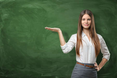 Beautiful young woman holding her open palm and showing at the empty area on the green chalkboard behind herの写真素材