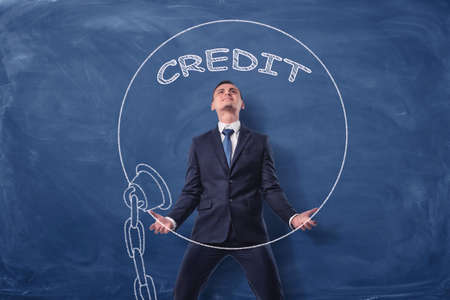 Businessman on blue chalkboard background lifting a big iron ball drawn in white with a word Creditの写真素材