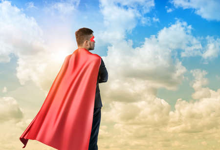 A businessman in superman cape standing turned back on the sky backgroundの写真素材