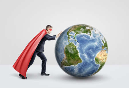 A businessman in a superhero red cape pushing and rolling a large globe on white background.の写真素材
