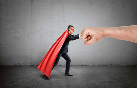 A businessman in a red cape and a mask pushing at a giant human fist on concrete background.の写真素材