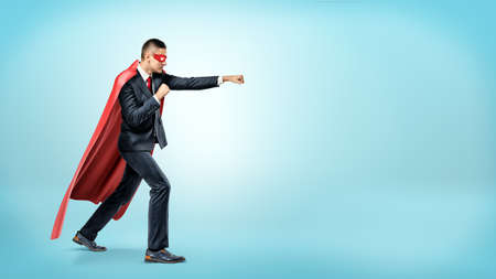 A businessman in a flowing red superhero cape and a mask throwing punches at an invisible enemy on blue background.の写真素材