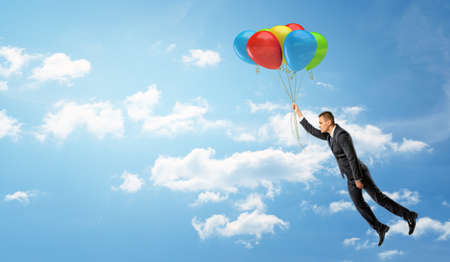 A businessman holding a batch of colorful balloons that let him fly through the clouds.の写真素材