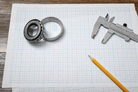 A vernier caliper holding a bearing, a pencil and a pair of compasses lying over drafting paper on wood background.の写真素材