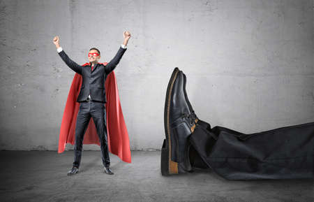 A smiling businessman in a superhero red cape with hands raised in success motion standing beside a giant human foot.の写真素材