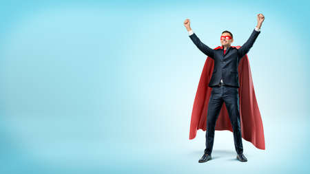A happy businessman in a superhero red cape standing in victory pose on blue background.