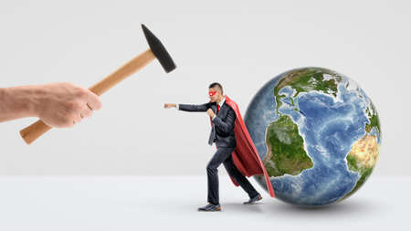 A businessman in a red superhero cape protecting a small globe behind him and punching a giant hand holding a hammer.の写真素材