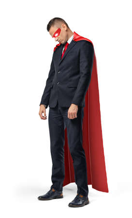 A sad businessman in a superhero red cape standing with his shoulders slumped and looking down.の写真素材