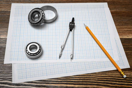 A vernier caliper holding a bearing, a pencil and a pair of compasses lying over drafting paper on wood background.の写真素材