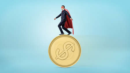 A businessman in a red superhero cape balancing on the edge of a giant golden coin with a USD sign.の写真素材