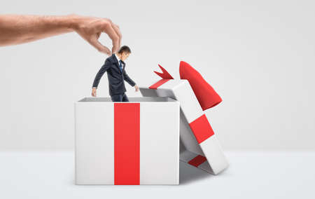 A large male hand getting a tiny businessman out from a white gift box with a red bow on white background.の写真素材