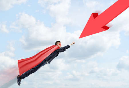 A businessman in a red cape and a mask flying through the clouds towards a huge red arrow. Problem solving. Business development. Goals and achievement.の写真素材