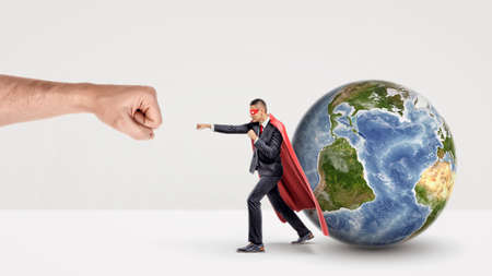 A small businessman in a red cape and a mask punching a giant fist with a small earth sphere behind him.の写真素材