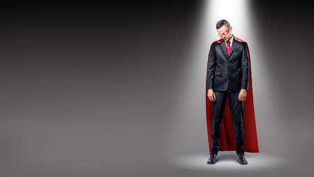 A sad businessman wearing a red superman cape standing in the spotlight with his shoulders slumped.の写真素材