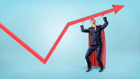 A businessman in a superman red cape supporting a red statistic arrow with his hands.の写真素材