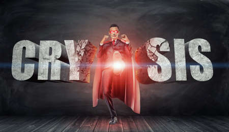 A businessman in a red cape and a mask kicking concrete letters reading Crysis with his foot.の写真素材