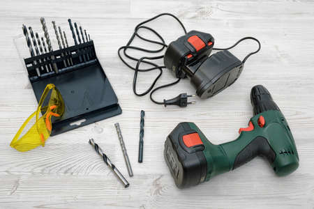 A cordless drill, a spare battery, a set of bits and yellow safety glasses on wooden table background.の写真素材