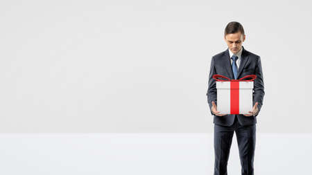 A businessman holding a gift box with a red bow in his arms looking down at it.