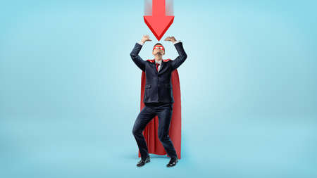 A businessman in a red cape and a mask cowering under a large red arrow pointing down at him.の写真素材