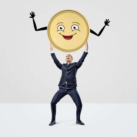 A happy smiling businessman holding a large golden coin with a smiley face over his head.の写真素材