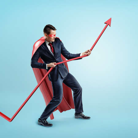 A businessman wearing a red superhero cape trying to hold a red statistic arrow with force on blue background.の写真素材