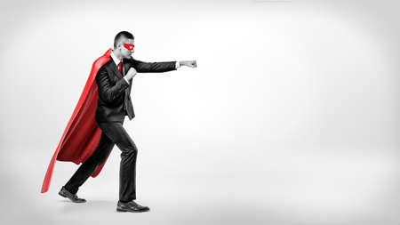 A businessman in a flowing red superhero cape and a mask throwing punches at an invisible enemy on white background.の写真素材