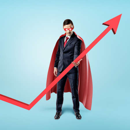 A businessman in a red flowing cape trying to keep the upward looking red arrow with his hand.の写真素材