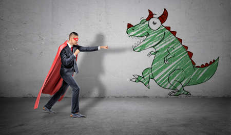 A businessman in a red cape standing in fighting pose ready to fight a picture of a dragon on the nearest wall.の写真素材