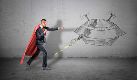 A businessman in a red superhero cape throwing punches at a wall drawing of an UFO striking at him.の写真素材